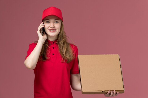 female courier in red uniform holding food box talking on phone on pink, uniform service delivery worker job