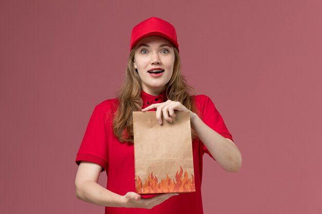 female courier in red uniform holding food package on light-pink, job uniform service worker delivery