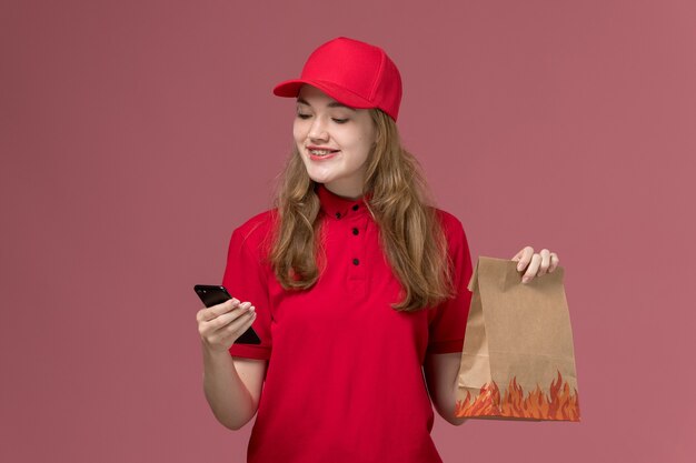 淡いピンクの電話を使用して笑顔の食品パッケージを保持している赤い制服を着た女性の宅配便、仕事の制服サービスワーカーの配達