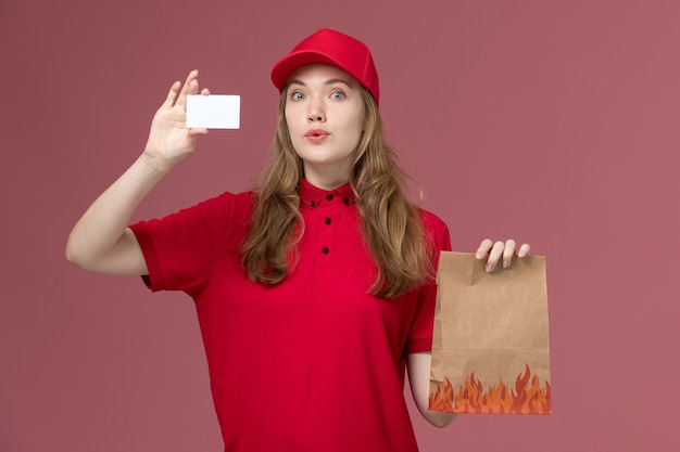 食品パッケージと淡いピンクの白いカードを保持している赤い制服を着た女性の宅配便、仕事の制服のサービスワーカーの配達