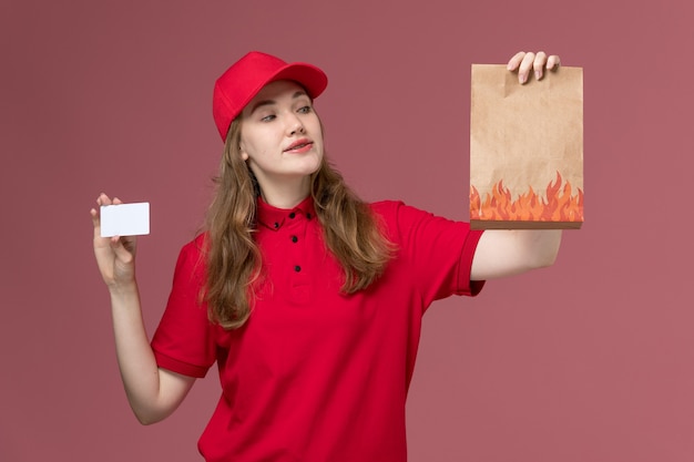 食品パッケージと淡いピンクの白いカードを保持している赤い制服を着た女性の宅配便、仕事の制服のサービスワーカーの配達