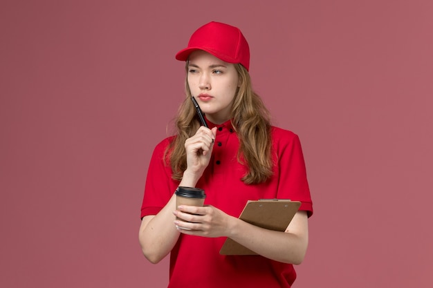 female courier in red uniform holding notepad and coffee cup with thinking expression on pink, uniform service delivery worker job