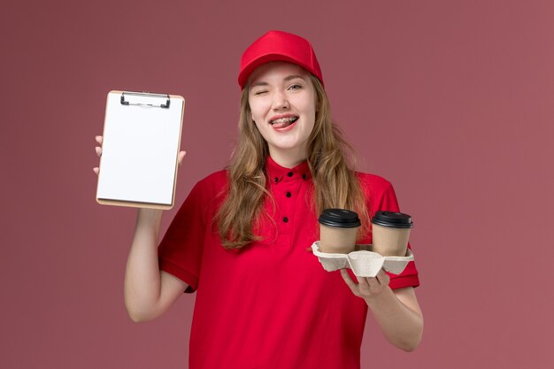 женщина-курьер в красной форме держит кофейные чашки блокнота на розовом, единообразный работник службы доставки