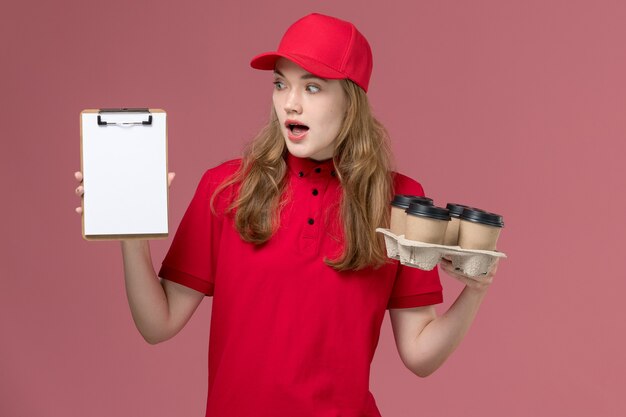 Foto gratuita corriere femminile in uniforme rossa che tiene il blocco note e le tazze di caffè sul lavoro rosa, uniforme dell'operaio di consegna del servizio