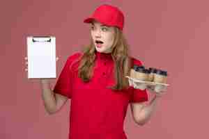 Free photo female courier in red uniform holding notepad and coffee cups on pink, uniform service delivery worker job