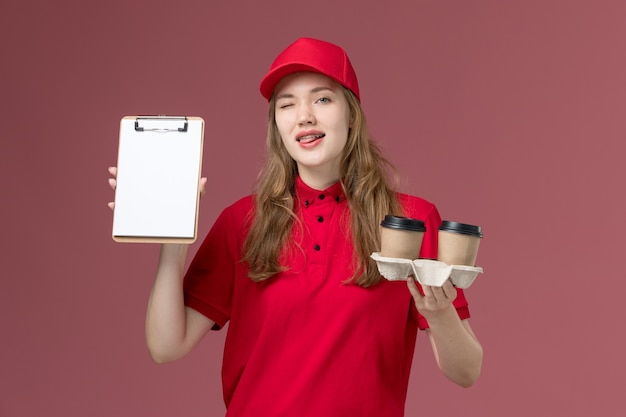 female courier in red uniform holding notepad and coffee on pink, uniform service delivering worker