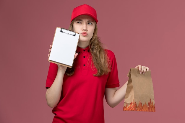 female courier in red uniform holding notepad and food package thinking on pink, uniform service delivery job worker