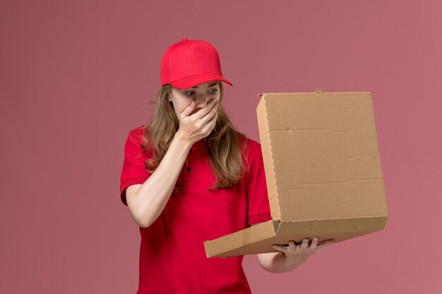 female courier in red uniform holding opening food box on light pink, job uniform service worker delivery