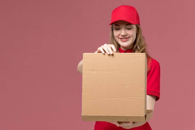 female courier in red uniform holding opening food box with smile on pink, uniform service delivering worker