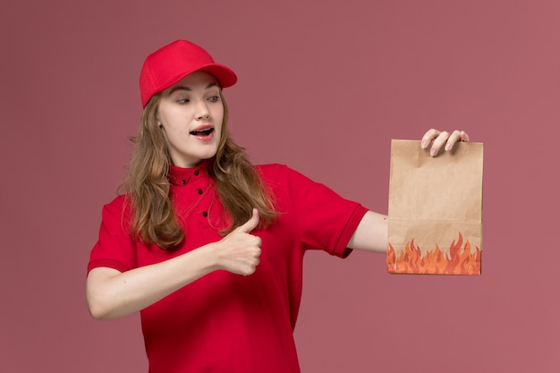 female courier in red uniform holding paper food package on the pink, uniform worker service delivery