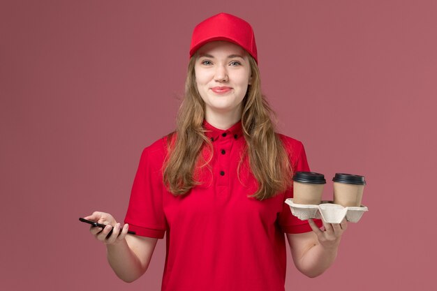 женщина-курьер в красной униформе держит смартфон и кофейные чашки на розовом, униформенный работник службы доставки