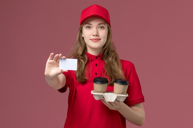 женщина-курьер в красной форме держит белую карточку и кофейные чашки с улыбкой на розовой униформе