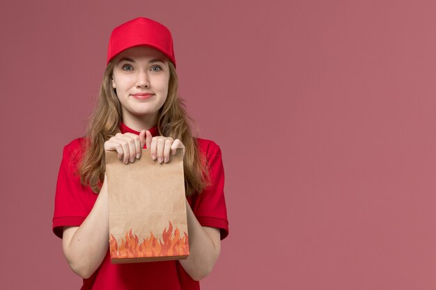 female courier in red uniform smiling holding food package on light-pink, job uniform service worker delivery