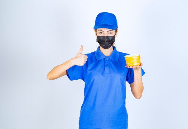 Female courier of the restaurant in blue uniform and face mask delivering a takeaway food and guaranteeing the good taste . 