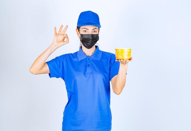 Female courier of the restaurant in blue uniform and face mask delivering a takeaway food and guaranteeing the good taste . 