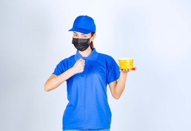 Female courier of the restaurant in blue uniform and face mask delivering a takeaway food and guaranteeing the good taste . 