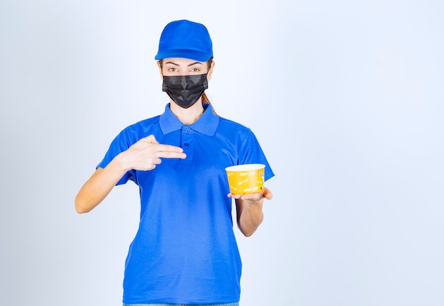 Female courier of the restaurant in blue uniform and face mask holding a takeaway food . 