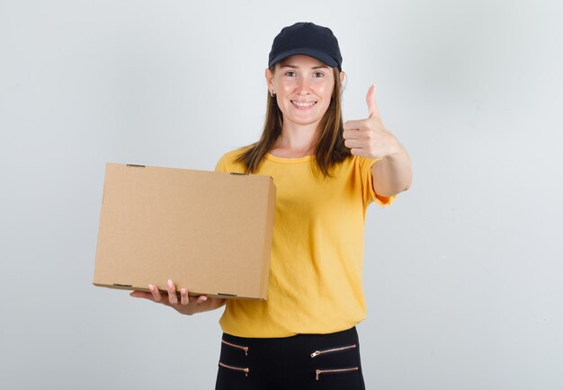 Female courier in t-shirt, pants, cap holding cardboard box with thumb up and looking glad