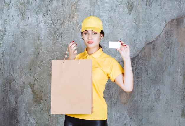 Бесплатное фото Курьер-женщина в желтой форме доставляет картонную сумку для покупок и показывает свою визитную карточку