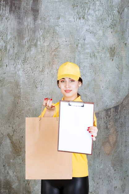 黄色いユニフォームを着た女性の宅配便業者が段ボールの買い物袋を配達し、署名のために空白を提示します。