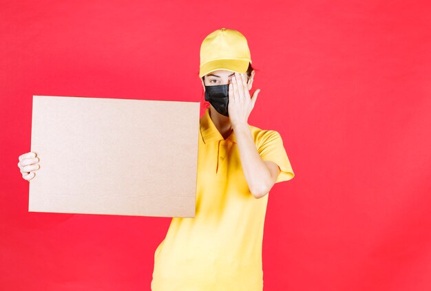 Female courier in yellow uniform and black mask holding the cardboard box and closing one eye