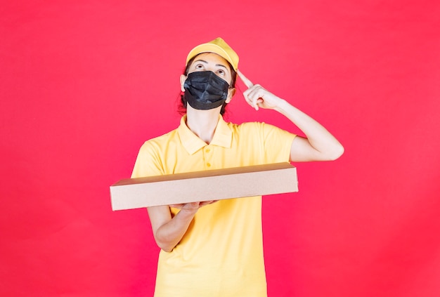 Free photo female courier in yellow uniform and black mask holding the cardboard box looks confused and thoughtful