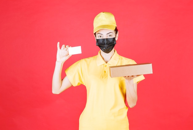 Female courier in yellow uniform and black mask holding the cardboard box and presenting her business card