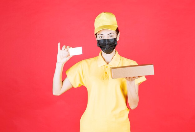 Женский курьер в желтой форме и черной маске держит картонную коробку и представляет свою визитную карточку