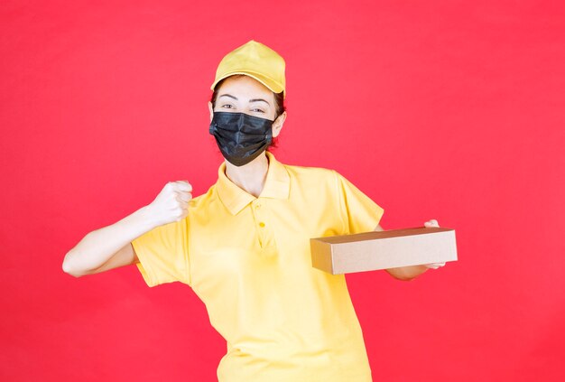 Female courier in yellow uniform and black mask holding the cardboard box and showing her fist