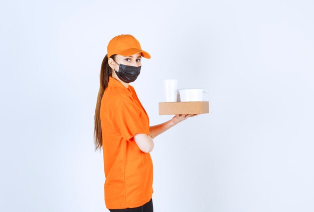 Female courier in yellow uniform and black mask holding a cardboard parcel, takeaway food and drink.