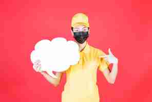 Free photo female courier in yellow uniform and black mask holding a cloud shape info board and pointing at it