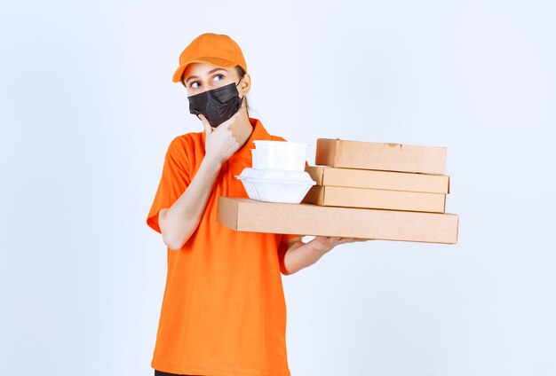 Female courier in yellow uniform and black mask holding multiple cardboard parcels and takeaway boxes and looks confused and thoughtful.