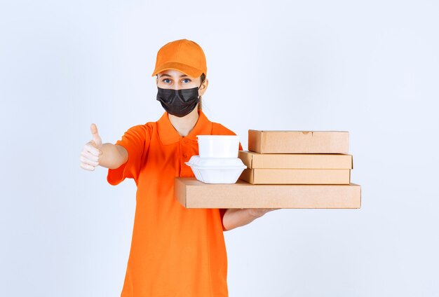 Курьер-женщина в желтой форме и черной маске держит несколько картонных посылок и коробок для еды на вынос, указывая на что-то.