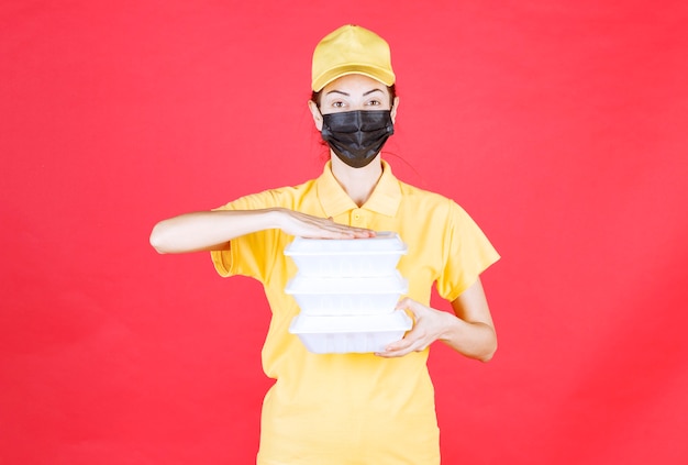 Female courier in yellow uniform and black mask holding multiple takeaway packages