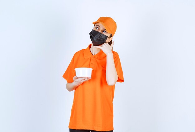 Female courier in yellow uniform and black mask holding a plastic cup and asking for a call