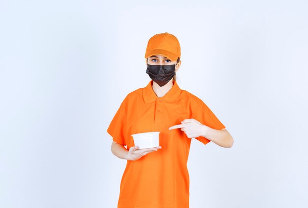 Female courier in yellow uniform and black mask holding a plastic cup and pointing at something.