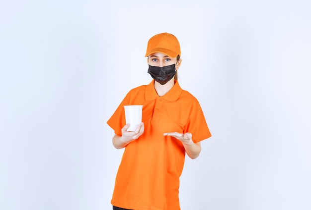 Female courier in yellow uniform and black mask holding a plastic takeaway cup and pointing at it.