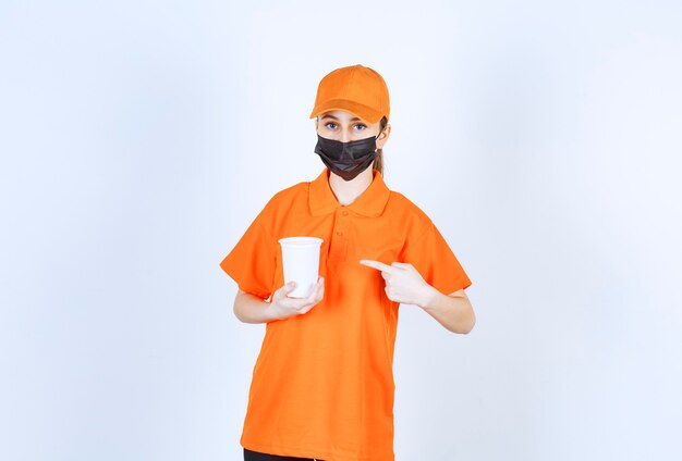 Female courier in yellow uniform and black mask holding a plastic takeaway cup and pointing at it.