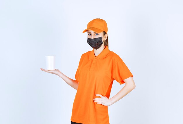 プラスチック製の持ち帰り用カップを保持している黄色の制服と黒のマスクの女性の宅配便。