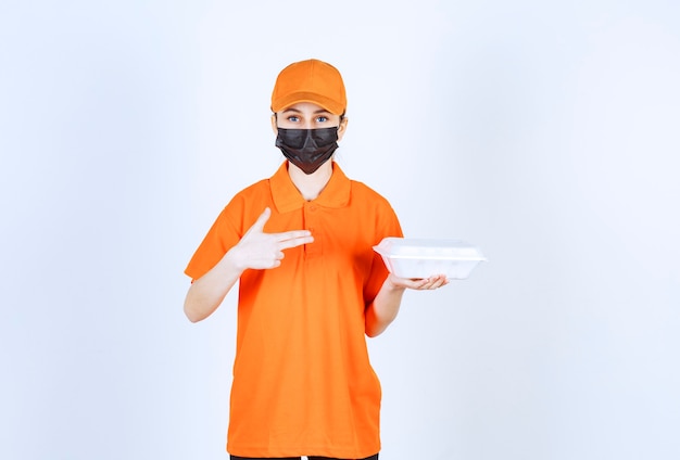 Female courier in yellow uniform and black mask holding a plastic takeaway food box and pointing at it.