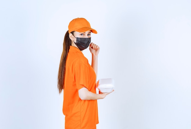 Free photo female courier in yellow uniform and black mask holding a plastic takeaway food box and showing her fist