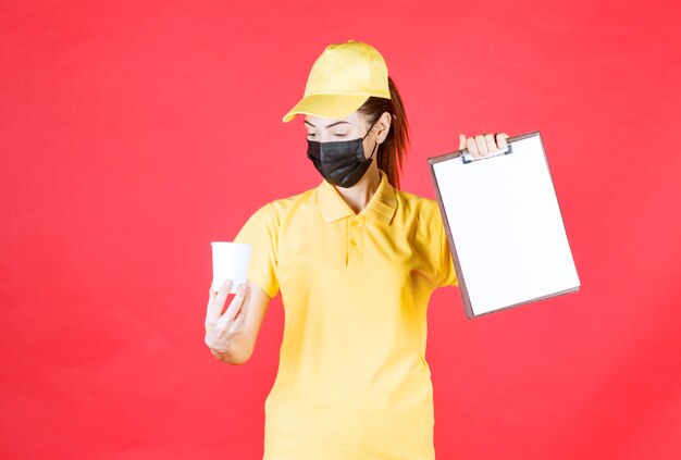 Female courier in yellow uniform and black mask holding a takeaway cup and a customer list