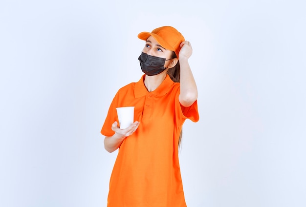 Female courier in yellow uniform and black mask holding a takeaway drink and thinking