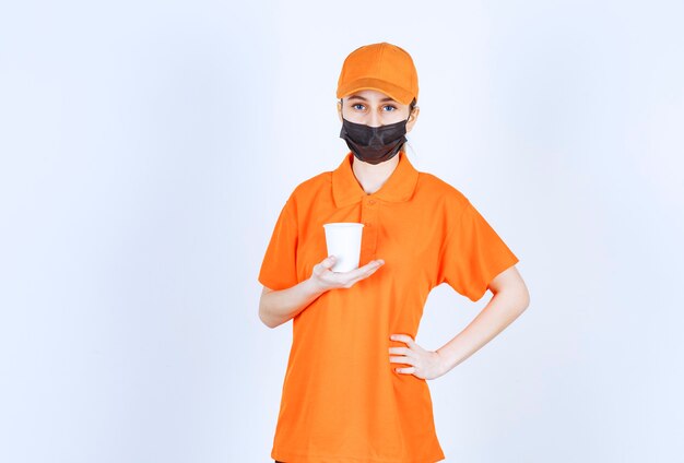 Female courier in yellow uniform and black mask holding a takeaway drink.