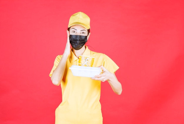 Female courier in yellow uniform and black mask holding a takeaway package and looks confused and thoughtful