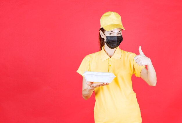 Female courier in yellow uniform and black mask holding a takeaway package and showing positive hand sign
