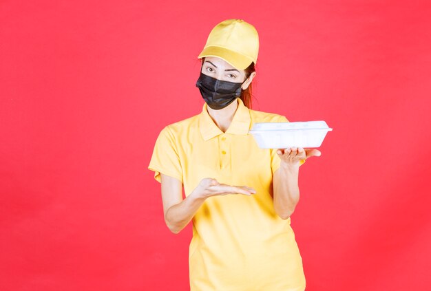 Female courier in yellow uniform and black mask holding a takeaway package