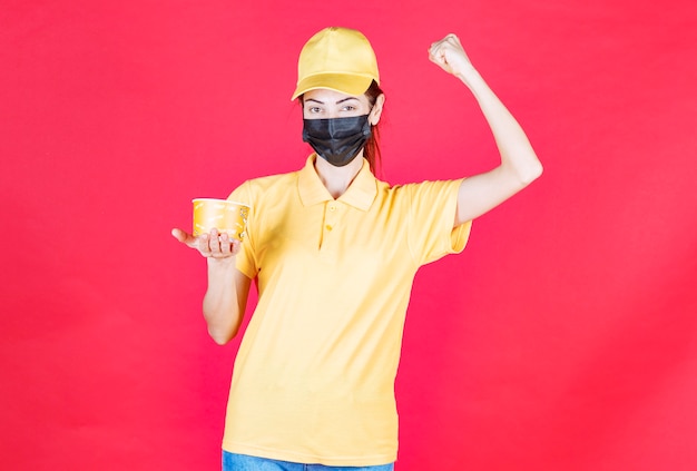 Free photo female courier in yellow uniform and black mask is delivering a noodles cup and showing her fist