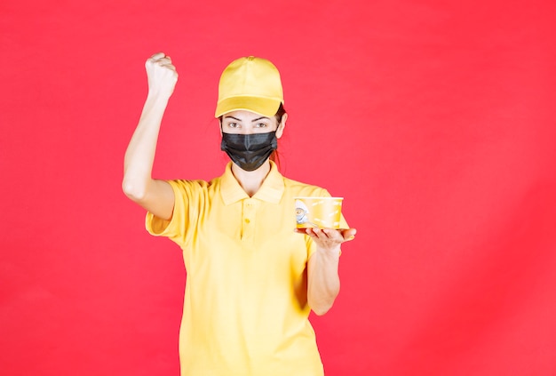Female courier in yellow uniform and black mask is delivering a noodles cup and showing her fist