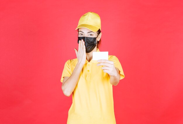 Female courier in yellow uniform and black mask presenting her business card and looks thoughtful and confused